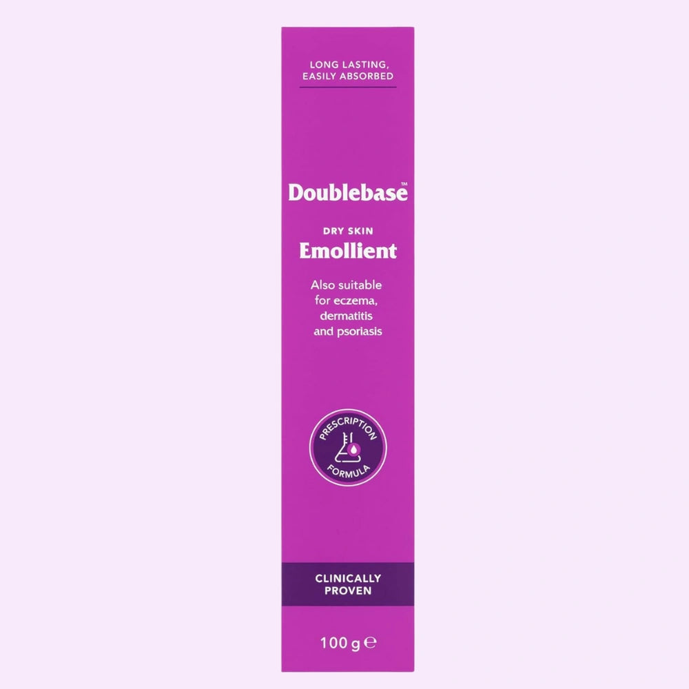 A tall magenta-purple tube of Doublebase Dry Skin Emollient 100g, shown vertically against a very pale lavender background. The tube features white text and a circular purple 'Prescription Only' seal.