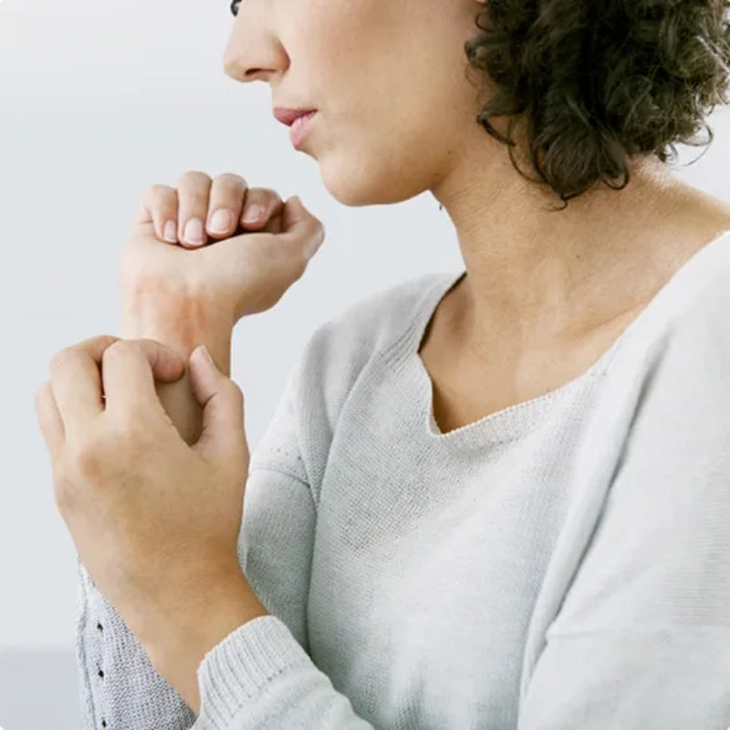 A close-up lifestyle shot of a woman in a light gray knit sweater holding and examining her wrist, shown from the side against a clean white background.