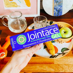 Hand holding a box of Jointace Aromatherapy Oils Gel on a table with a plate of fruit and a glass of water.
