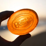A hand holds a translucent amber-orange oval bar of Pears soap against a bright, sunlit background. The embossed brand name 'Pears' is clearly visible on the soap surface.
